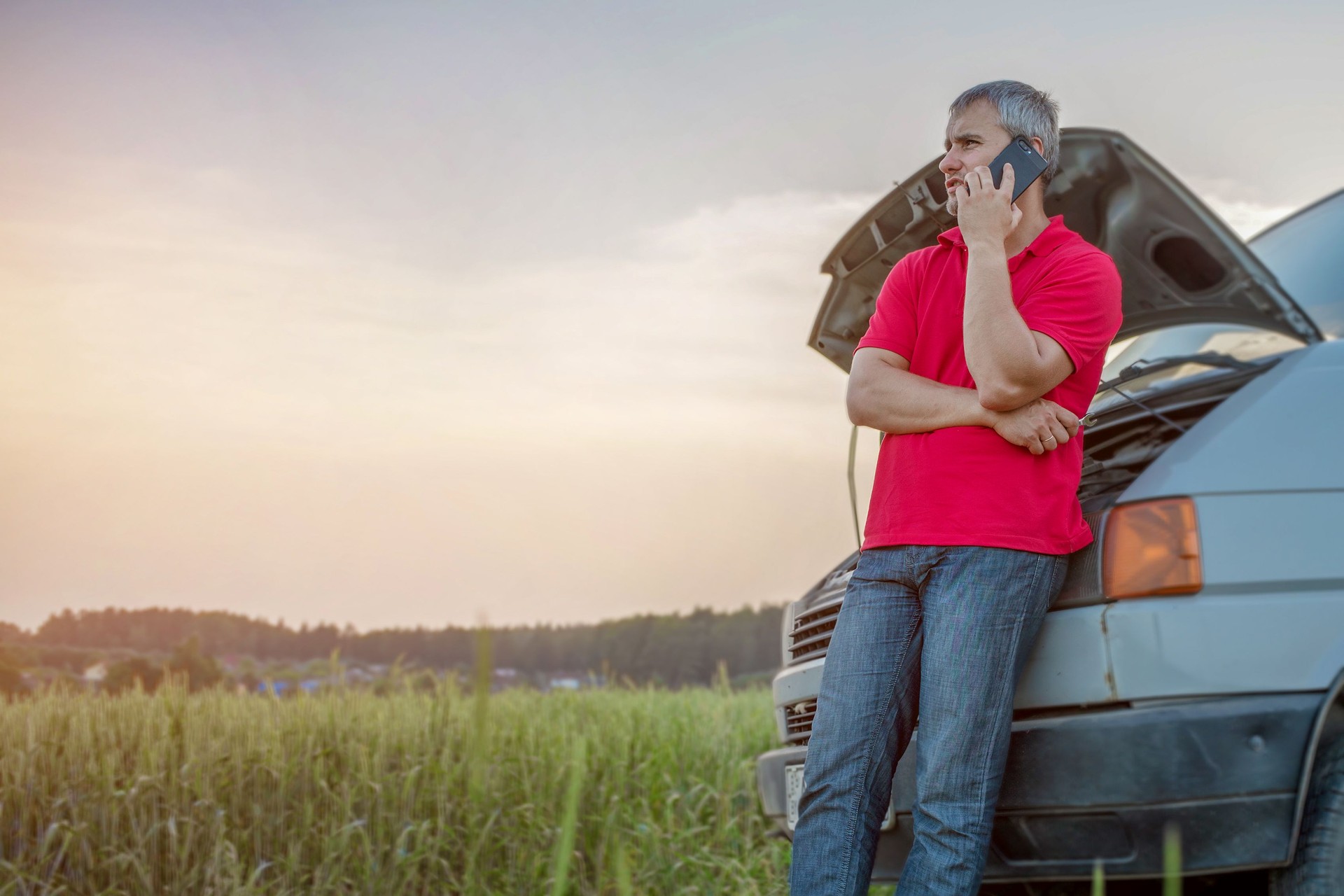 Man talking by phone leaning to van standing on rural road with raised hood